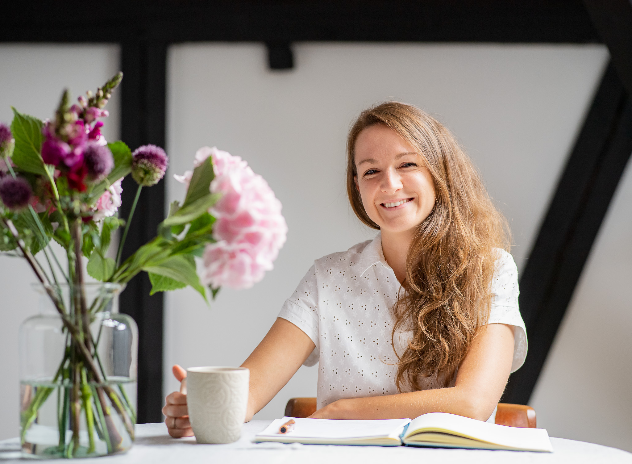Vanessa Büchter, Coach für integrative Darmgesundheit, sitzt am Tisch, lächelt in die Kamera, mit Notizbuch und Tasse.